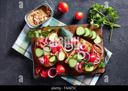 Cod Leber Creme auf Toast mit Gurken, Tomaten und roter Zwiebel Ringe auf einem Schneidebrett mit Zutaten auf einer konkreten Tabelle, Ansicht von oben, flatla Stockfoto