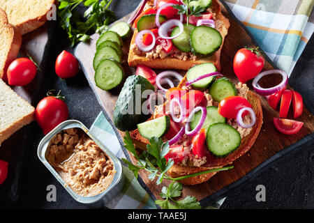 In der Nähe von Toast mit Lebertran Pastete, mit geschnittenen Gurken, Tomaten und roter Zwiebel Ringe auf einem Schneidebrett mit Zutaten auf einer konkreten ta gekrönt Stockfoto