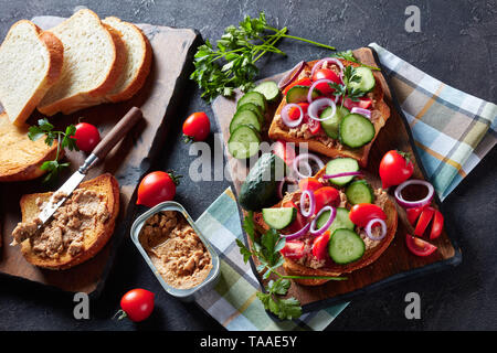 Knuspriges Weißbrot, Toast mit Lebertran Pastete, mit geschnittenen Gurken, Tomaten und roter Zwiebel Ringe auf einem Schneidebrett mit Zutaten auf einem kitc gekrönt Stockfoto