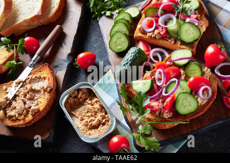 Toast für Sommer Frühstück mit Lebertran Pastete, gekrönt mit in Scheiben geschnittenen Gurken, Tomaten und roter Zwiebel Ringe auf einem Schneidebrett mit Zutaten aus einem Ki Stockfoto