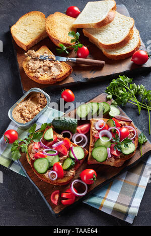 Toast für Sommer Frühstück mit Lebertran Pastete, gekrönt mit in Scheiben geschnittenen Gurken, Tomaten und roter Zwiebel Ringe auf einem Schneidebrett mit Zutaten aus einem Ki Stockfoto