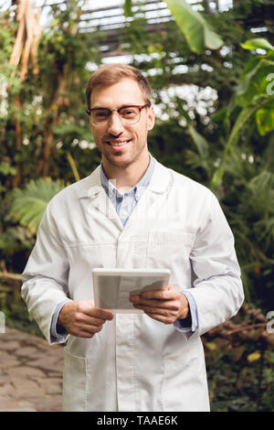 Lächelnd stattlichen Wissenschaftler im weißen Mantel und Gläser halten digitale Tablet in Orangerie Stockfoto