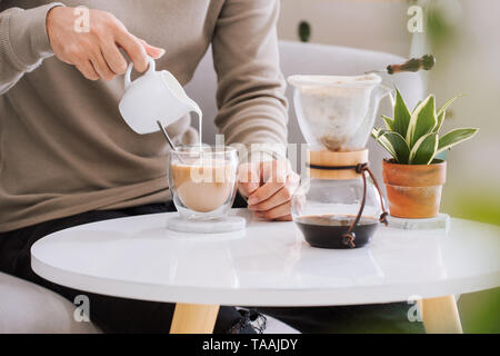 Kaffee trinken: des Menschen Hand gießt Milch in ein Glas Espresso in Wohnzimmer. Stockfoto