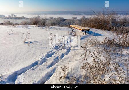 Winterlandschaft, sonnigen Tag. Spuren im Schnee führen zu einer Holzbank, die auf dem hohen Ufer des Flusses steht. Stockfoto