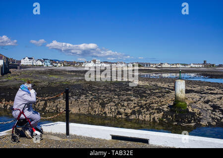 Ältere Frau tragen Steppjacke sitzen an der Seite des äußeren Hafenmauer in Castletown, Insel Man mit Blick auf die Bucht bei Ebbe Stockfoto