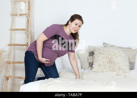 Schwangere europäische Frau mit Bauchschmerzen ihr Nelly und auf dem Bett sitzend Holding, müde und unzufrieden, eine Person Stockfoto