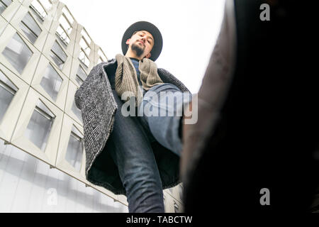 Porträt einer stilvollen hübscher junger Mann mit Mantel im Freien. Low Angle View von ein Mann mit einem Mantel und Schal auf der Suche überzeugt. Stockfoto
