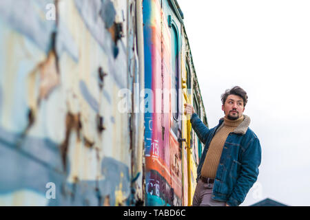 Porträt einer stilvollen hübscher junger Mann mit einem denim Wolljacke im Freien auf einen Zug. Ein Mann mit einem denim Wolljacke und einen Hut, mit Blick auf den Ca Stockfoto