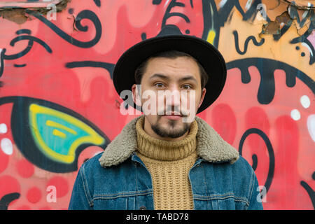 Porträt einer stilvollen hübscher junger Mann mit einem denim Wolljacke im Freien. Ein Mann mit einem denim Wolljacke und einem Hut, in die Kamera schaut. Stockfoto