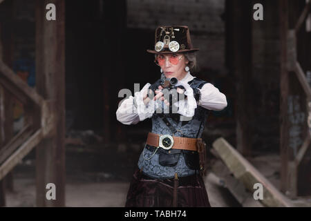 Ältere Dame in eine Steampunk Kostüm an einer verlassenen Fabrik mit der Waffe in der Hand. Stockfoto