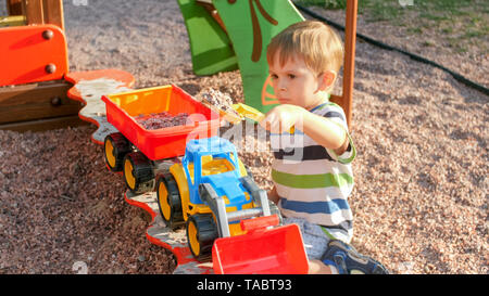 Portrait von freundlich lächelnden kleinen Jungen gießen Sand in Spielzeug-LKW mit Anhänger. Kinder spielen und sich auf dem Spielplatz im Park Stockfoto