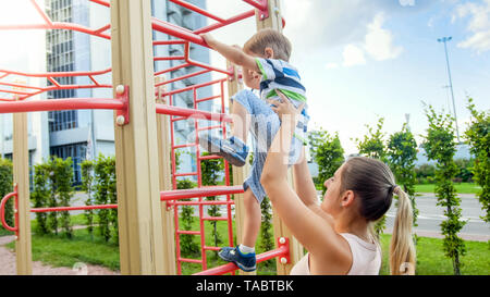 Süße 3 Jahre altes Kleinkind junge klettern hoch auf die Kinder playghround im Park, während seine Mutter unterstützt und ihn hält Stockfoto