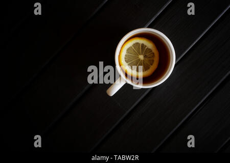 Eine weiße Tasse mit schwarzem Kaffee und einem Stück Zitrone steht auf einem dunklen Holztisch. Blick von oben. Minimalismus. Tageslicht. Stockfoto