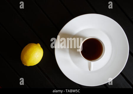 Eine weiße Tasse mit schwarzem Kaffee steht in einer Untertasse Neben einer gelben Zitrone auf einem dunklen Holztisch. Blick von oben. Minimalismus. Tageslicht. Stockfoto