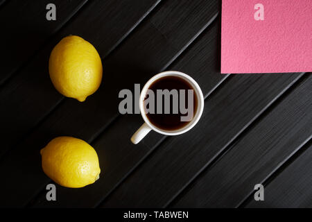 Eine weiße Tasse mit schwarzem Kaffee steht auf einem schwarzen Tisch neben zwei gelbe Zitronen und ein rosa Serviette. Blick von oben. Minimalismus. Tageslicht Stockfoto
