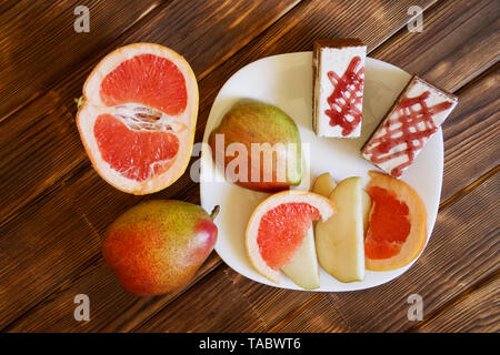 Kuchen und Scheiben von Grapefruit und Birnen liegen in einer weißen Platte auf einem Holztisch aus Kiefernholz Planken. Frühstücksbuffet im authentischen natürlichen Hotel. Tageslicht. Stockfoto