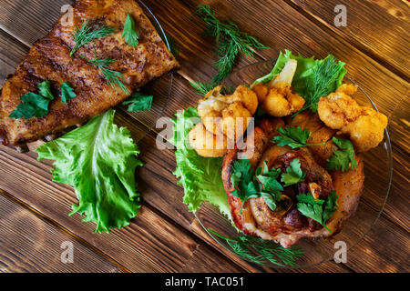 Köstliche gegrillte Rippchen und Knöchel, gewürzt mit Petersilie, Salat, Blumenkohl und Dill. Gebackenes Fleisch liegt auf einer hölzernen rustikalen Tisch aus natur Stockfoto