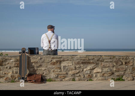 Rückansicht der Mann mit Laptop und Longboard sitzen auf einer Mauer vor der Strand und das Meer in der Ferne suchen Stockfoto