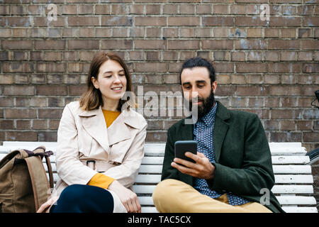 Paar auf einer Bank an eine Mauer, die gemeinsame Nutzung von Handy sitzen Stockfoto