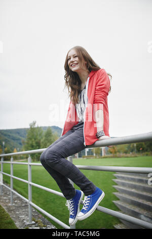 Glückliches junges Mädchen sitzt auf Zaun an einem Sportplatz Stockfoto