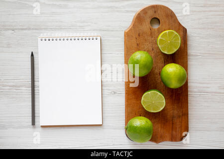 Ganzen und geschnittenen grünen Zitrusfrüchten Limes auf einer rustikalen Holzbrett, Notepad mit Bleistift über weiß Holz- Oberfläche, Ansicht von oben. Stockfoto