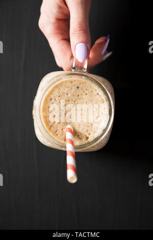 Woman's Hand mason Glas Glas Becher gefüllt mit Banane, Kiwi, Apfel Smoothie über schwarze Oberfläche, Ansicht von oben. Close-up. Stockfoto