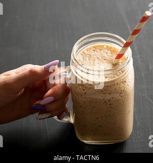 Woman's Hand mason Glas Glas Becher gefüllt mit Banane, Kiwi, Apfel Smoothie über schwarze Oberfläche, Low Angle View. Close-up. Stockfoto