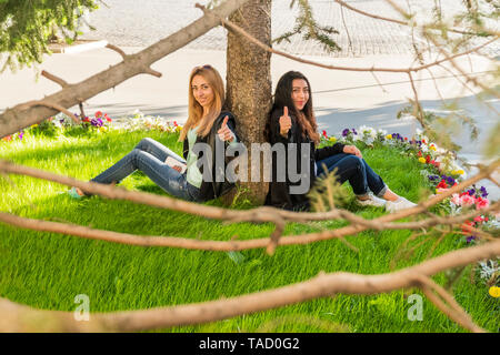 Zwei lächelnde Studierende aus verschiedenen Ländern sitzen auf dem Gras von einem Baum und mit Daumen nach oben an der Kamera Stockfoto