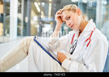 Junge Frau als Arzt oder Krankenschwester sitzt müde und erschöpft auf dem Boden Stockfoto
