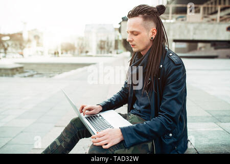 Junge modische kaukasischen Mann mit Dreadlocks in Leder Jacke sitzen auf der Treppe mit Laptop gekleidet. Remote funktioniert IT-Spezialist Stockfoto