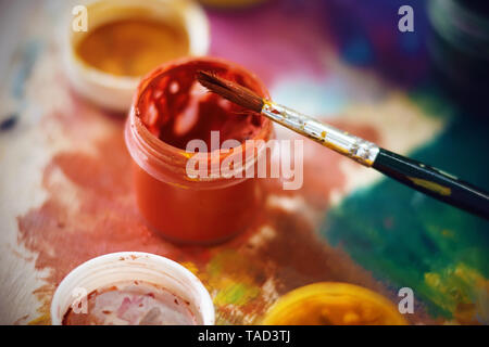 Ein Glas mit roter Gouache und einer Bürste, die für die Zeichnung verwendet wurde, sind auf der Palette, in unterschiedlichen Farben lackiert Stockfoto