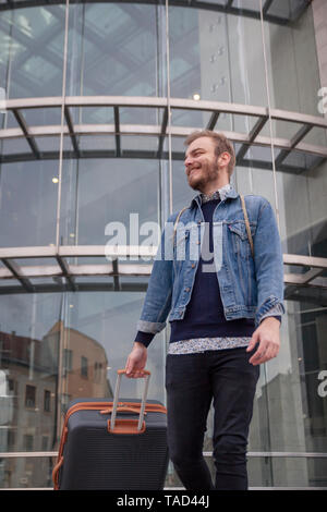 Aufnahme von unten, ein junger Mann - Traveler oder Tourist, lächelnd und suchen seitwärts, während seine Koffer tragen. Modernes Gebäude außen, Glaswand, Stockfoto