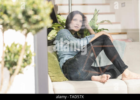 Nachdenkliche Frau auf der Couch hinter Fensterglas zu Hause sitzen Stockfoto