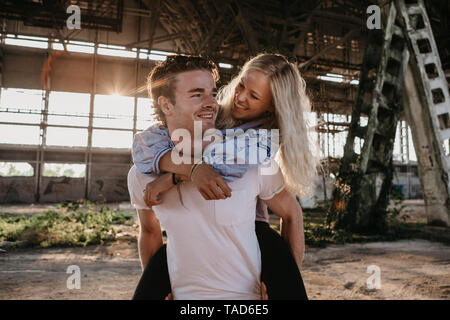 Glückliche junge Mann, Freundin ein piggyback Ride in einer alten Halle Stockfoto