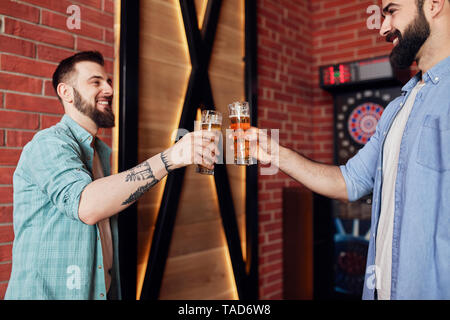 Zwei glückliche Freunde klirren Biergläser an der Dartscheibe in einem Pub Stockfoto