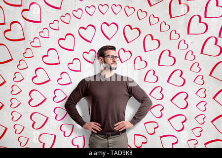 Hübscher junger Mann mit Brille, stehend, Hände in der Taille, Weg suchen, an der Wand mit kreisförmigen Muster von Hand gezeichnete Herzen. Stockfoto