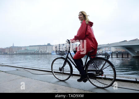 Dänemark, Kopenhagen, glückliche Frau Reiten Fahrrad an der Waterfront im regnerischen Wetter Stockfoto