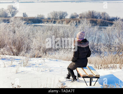 Eine junge Frau, die in den Strahlen der aufgehenden Sonne im Winter die Morgendämmerung am Ufer des südlichen Bug Fluss grüßt, sitzt auf einer Holzbank und schaut in den d Stockfoto