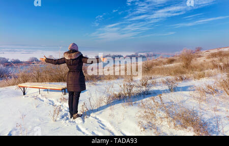 Eine junge Frau, die in den Strahlen der aufgehenden Sonne grüßt ein Winter im Morgengrauen auf der Bank des südlichen Bug River, auf einem Hügel in der Nähe einer Holzbank und Loo Stockfoto