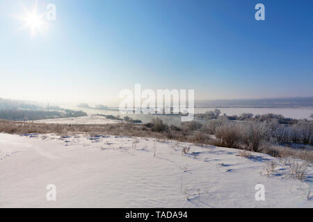 Ländliche Winterlandschaft, die Strahlen der Sonne in einem Dunst von blue sky liegen auf einer kurvenreichen Fluss und schneebedeckte Felder, Büsche und Bäume. Stockfoto