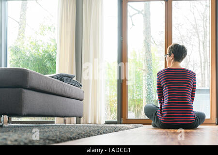 Rückansicht der Frau auf dem Boden sitzend Wohnzimmer Blick aus Fenster Stockfoto