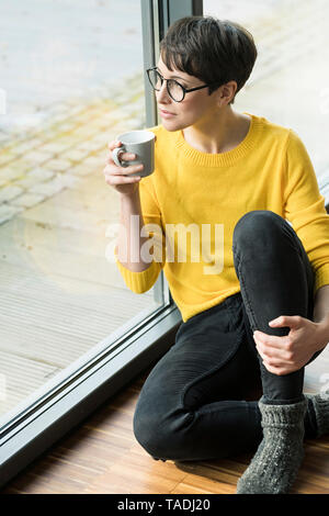 Frau mit Tasse Kaffee auf dem Boden sitzend zu Hause Blick aus Fenster Stockfoto