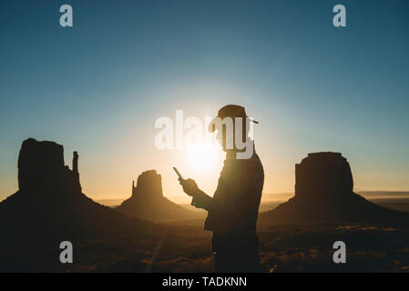 USA, Utah, Monument Valley, Silhouette der Mann mit Cowboyhut auf Handy bei Sonnenaufgang auf der Suche Stockfoto