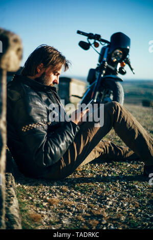 Mann mit custum Motorrad mit einer Unterbrechung auf dem Boden sitzend mit Handy Stockfoto