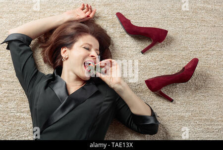 Müde Geschäftsfrau in schwarzem Kleid liegen auf dem Teppich und essen Erdbeeren. Stockfoto