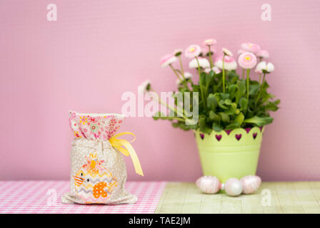 Eine kleine Tasche bestickt mit einem Küken wird mit einem kleinen Geschenk gefüllt. Es ist Ostern, wenig Ostereier liegen mit Frühling Blumen Bellis perennis auf Holz. Pi Stockfoto