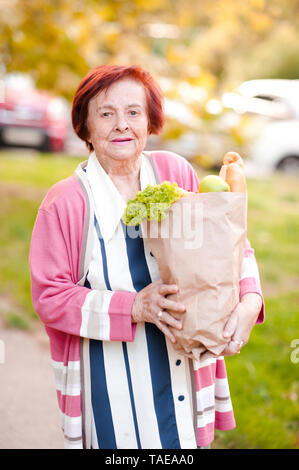 Lächelnd senior Frau 70-80 Jahre alten Holding Papiertüte mit Produkten im Freien. Mit Blick auf die Kamera. Stockfoto