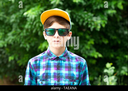 Stilvolle Teenager tragen Sonnenbrille, Mütze und trendiges Shirt im Freien. Mit Blick auf die Kamera. Posing über Natur Hintergrund. Stockfoto