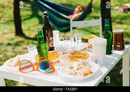 Tabelle mit Flaschen Bier und Essen auf Sommer Garden Party - die Frau, die in einer Hängematte im Hintergrund Stockfoto