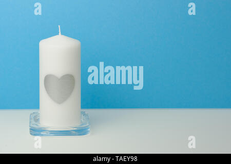 Weiße große Kerze mit einem silbernen Herz auf ihn vor einem blauen Hintergrund auf einem weißen Tisch, mit Kopie Raum für Grußkarten Stockfoto